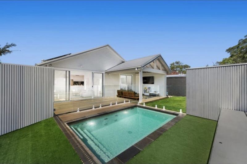 White weatherboard home with a deck, lawn and in-ground pool under a blue sky